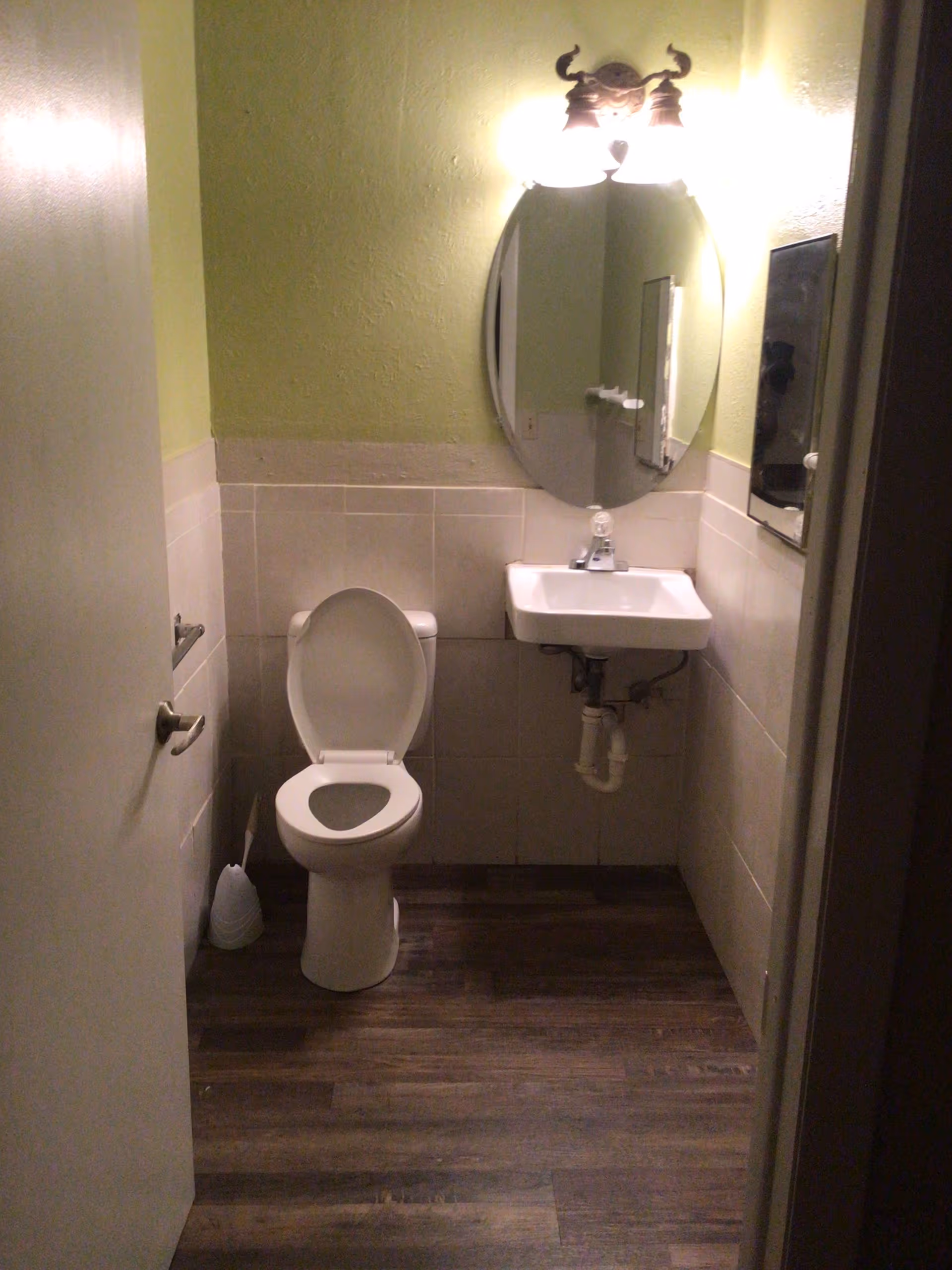 Small bathroom with a white toilet with the lid open, a white wall-mounted sink, an oval mirror above the sink, a wall light fixture with two bulbs above the mirror, light green walls, beige tiles halfway up the walls, and dark wood flooring. A toilet brush is placed on the floor next to the toilet.
