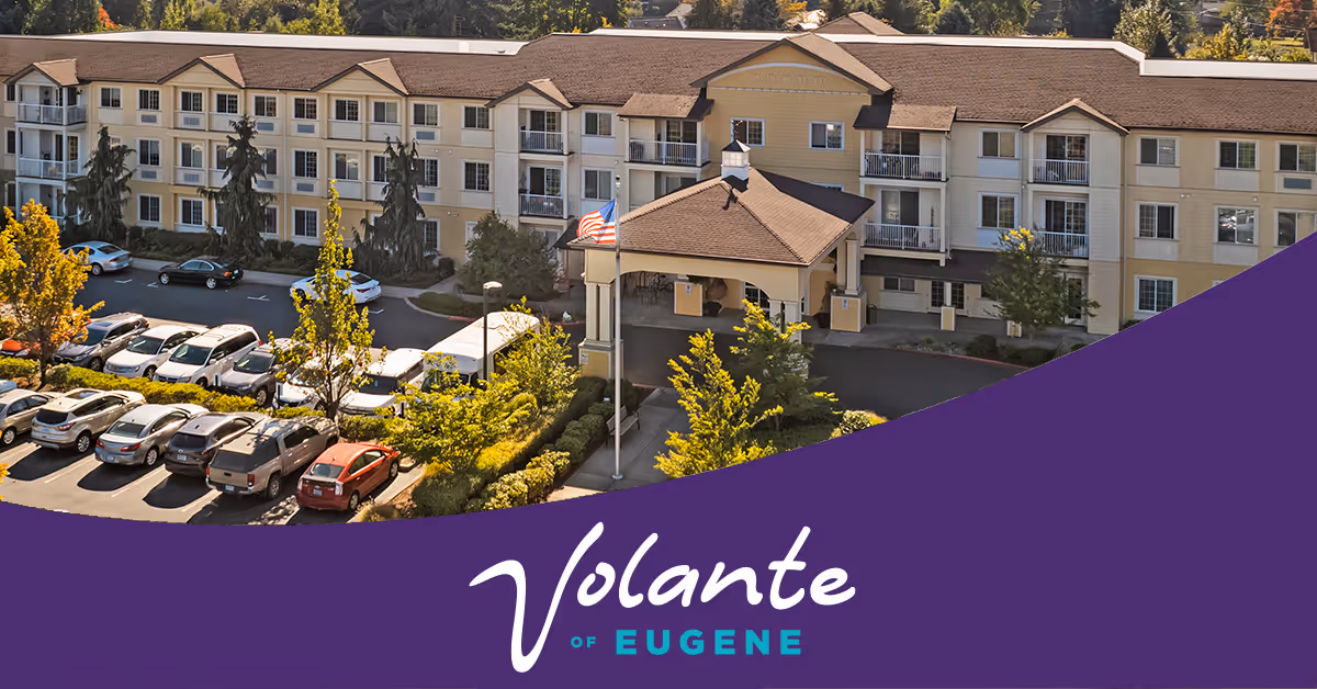 Exterior view of a three-story senior living facility building with a covered entrance, multiple balconies, and a parking lot with several cars. Trees and landscaping surround the building.