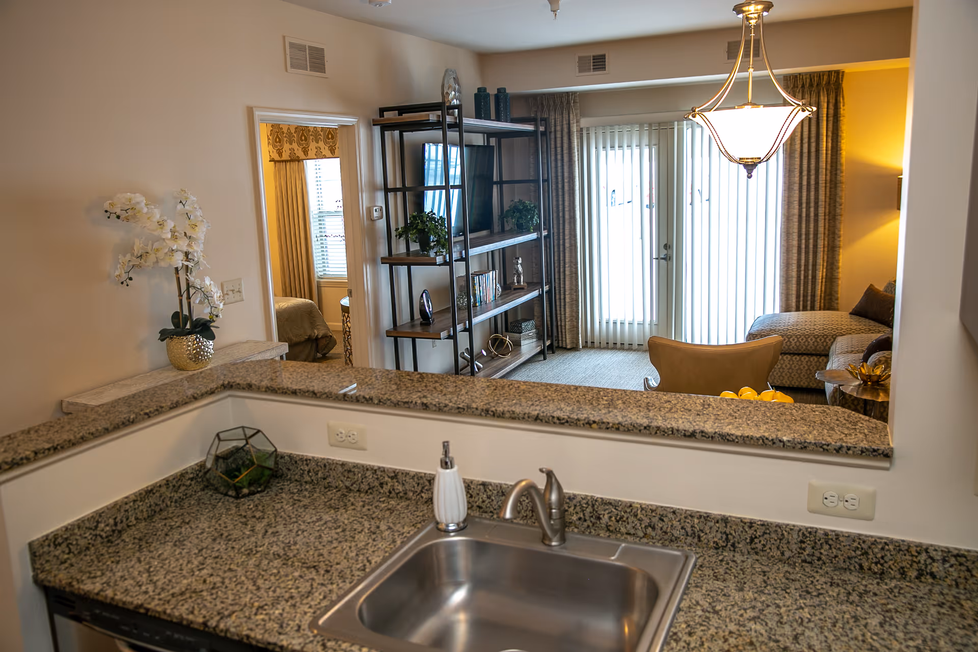 View from a kitchen sink and granite countertop into a living room with a bookshelf, sliding glass doors and seating.