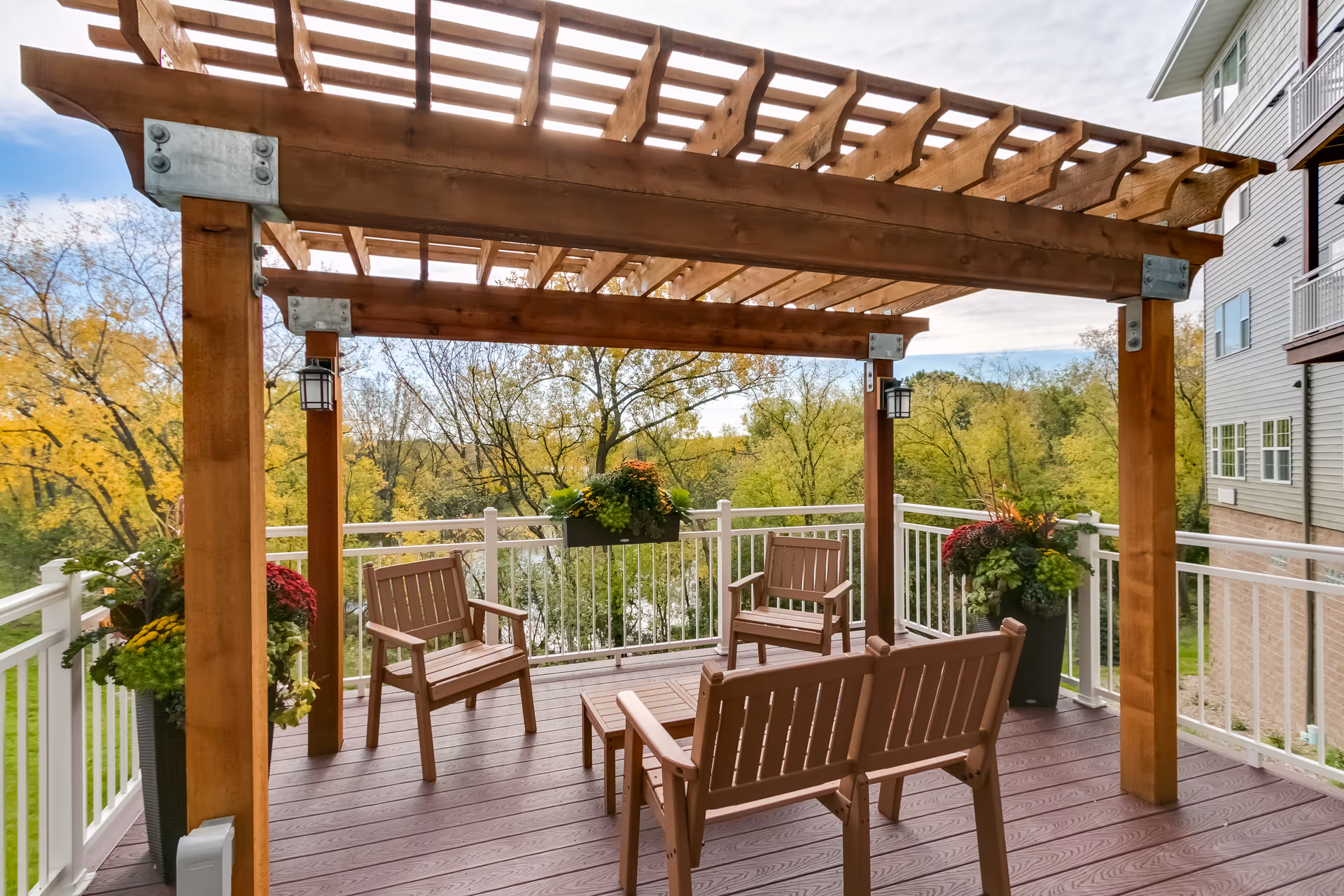 Outdoor patio area with a wooden pergola overhead, surrounded by white railings. The patio has wooden chairs and a bench arranged around a small table. There are large planters with colorful flowers and greenery on the corners. Trees with autumn foliage are visible in the background.
