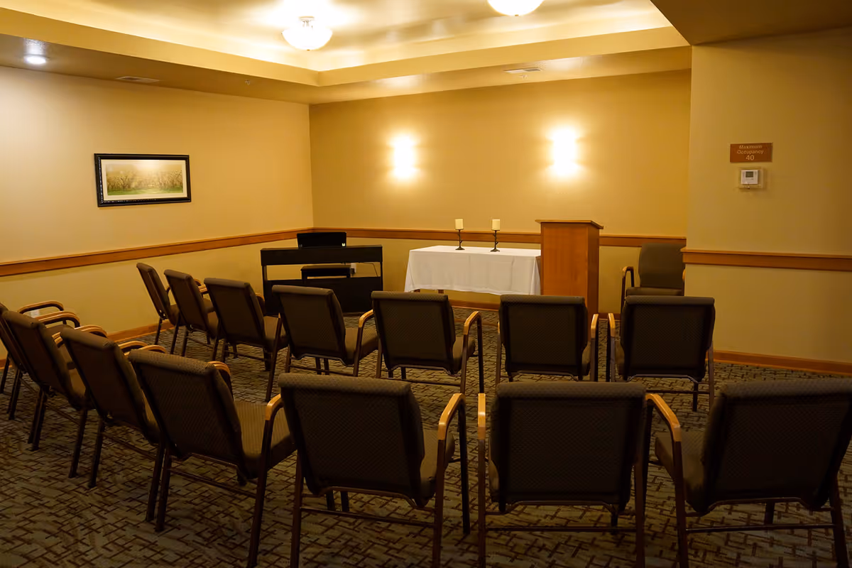 A small meeting or chapel room with rows of chairs facing a table covered with a white cloth, two candles, a wooden podium, and an electronic keyboard against the wall. The room has beige walls, carpeted floor, soft lighting, and a framed picture on one wall.