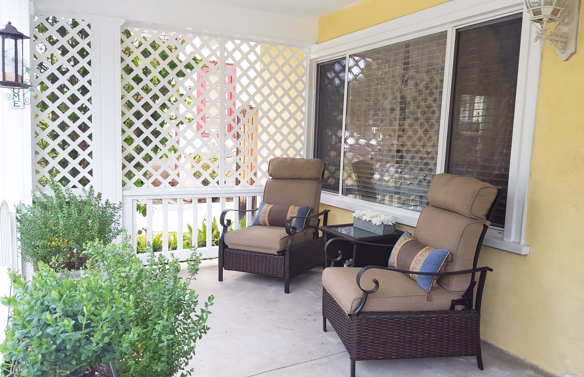 A cozy outdoor patio area with two cushioned wicker armchairs, each with decorative pillows. Between the chairs is a small table with a flower arrangement. The patio has white lattice panels and railings, with green plants in pots along the edge. The wall behind the chairs is yellow with a large window and a decorative wall lantern.