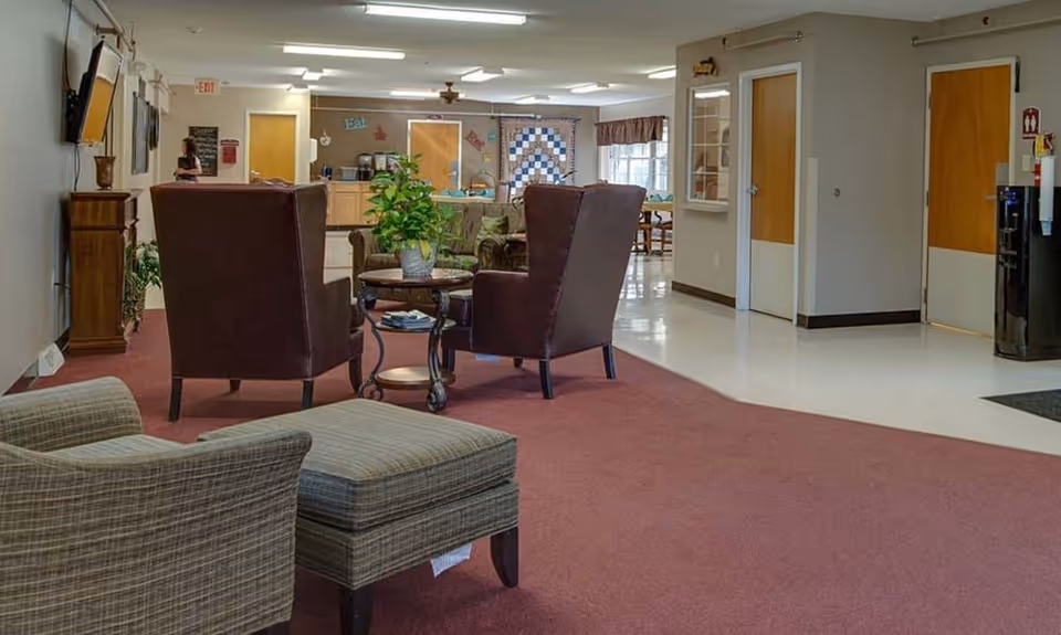 A carpeted common lounge area with armchairs and a small table opening into a dining/kitchen area.