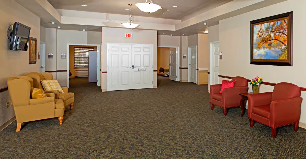 A spacious interior common area with beige walls and carpeted floor. There are two yellow armchairs with pillows on the left side and two red armchairs with a small table and a flower vase on the right side. A large framed painting of a tree with autumn leaves hangs on the right wall. A flat-screen TV is mounted on the left wall. Several doors and an exit sign are visible in the background under ceiling lights.
