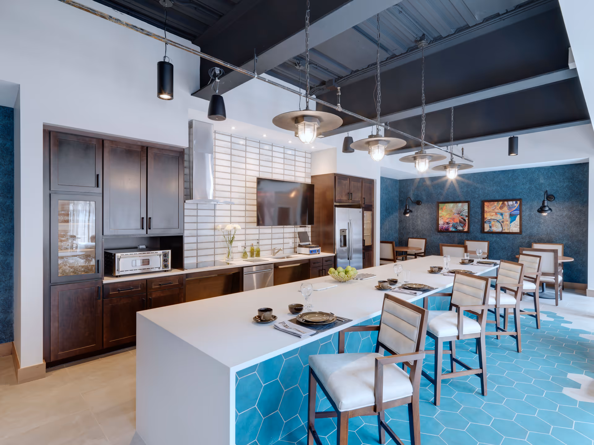 Modern kitchen and dining area with a long white island counter featuring place settings and chairs. The kitchen has dark wood cabinets, stainless steel appliances, a mounted flat-screen TV, and a white tiled backsplash. The floor has blue hexagonal tiles near the island, and the walls are painted blue with two colorful framed artworks. Pendant lights hang from a dark ceiling above the island.
