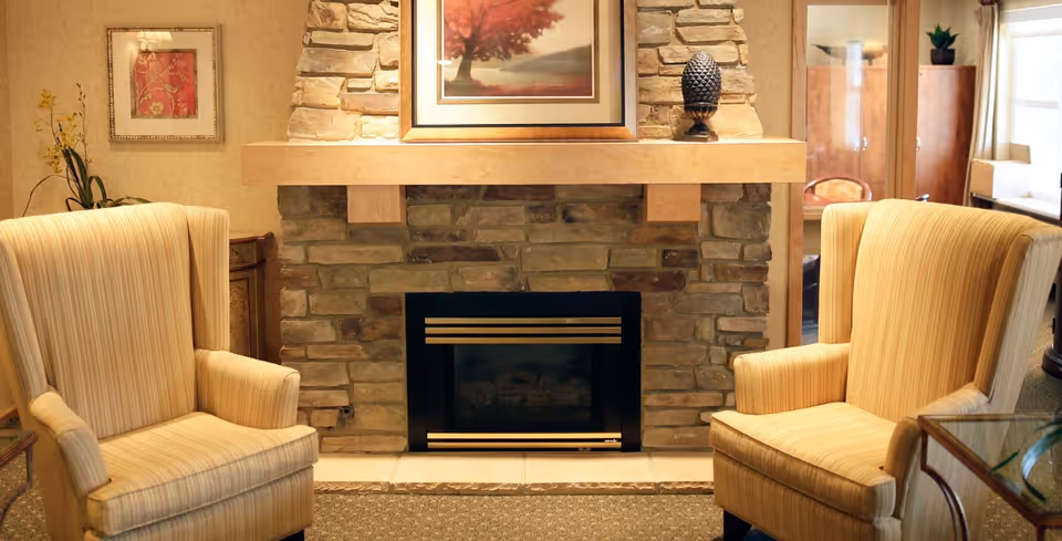 A cozy sitting area with two beige striped armchairs facing a stone fireplace with a wooden mantel. Above the mantel is a framed picture of a tree with red leaves. The room has warm lighting and traditional decor, including a small table with a plant and a cabinet with a mirror in the background.
