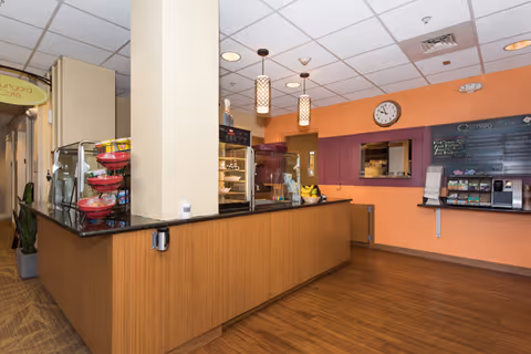 Interior view of a small cafeteria-style counter with display cases, pendant lights, a clock, and a menu board.