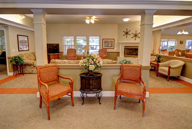 Communal lounge area with armchairs, a small decorative table topped with a floral arrangement, and additional seating around a fireplace.