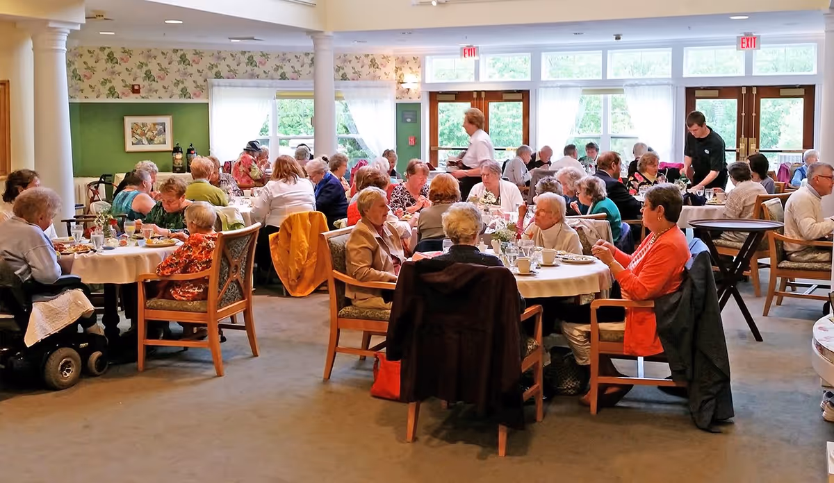 A large group of elderly residents seated at round tables in a bright assisted-living dining room during a meal.