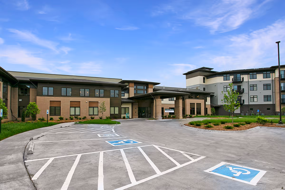 Exterior view of Prairie Gate senior living facility showing a modern multi-story building with large windows, a covered entrance, and a parking lot with designated handicapped parking spaces under a partly cloudy blue sky.