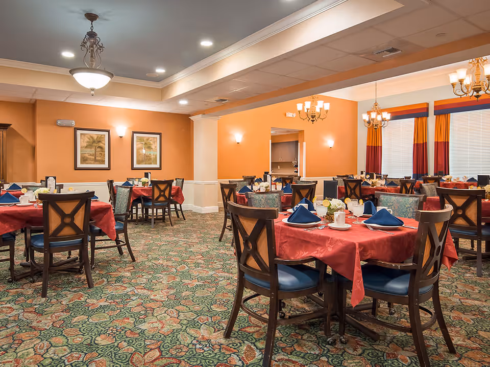 Bright dining room with round tables covered in red tablecloths, blue napkins, and wooden chairs.