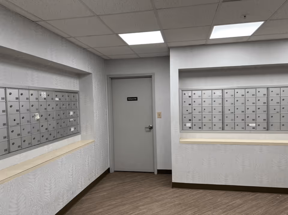Interior view of a mailroom area with two sets of metal mailboxes mounted on opposite walls. There is a gray door labeled 'Employees Only' in the center, and the room has a drop ceiling with fluorescent lighting and wood-patterned flooring.