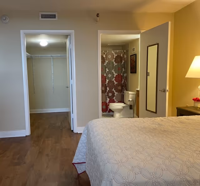 A bedroom with a bed in the foreground, a nightstand lamp, an open closet doorway on the left and an open bathroom with a toilet and patterned shower curtain on the right.