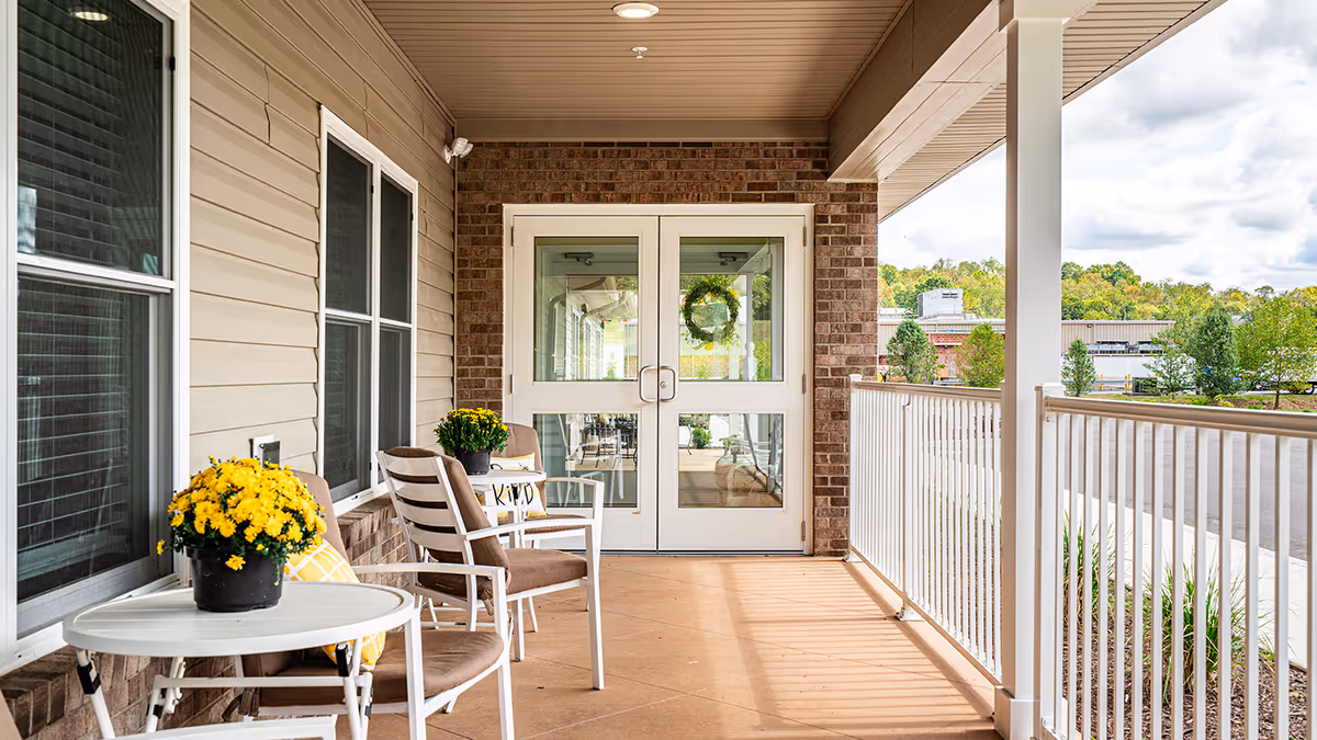 Covered porch area with two brown cushioned chairs and a small white round table holding a pot of yellow flowers. The porch has beige siding and brick walls, white railing, and double glass doors with a wreath visible inside. Trees and a parking lot are visible in the background.