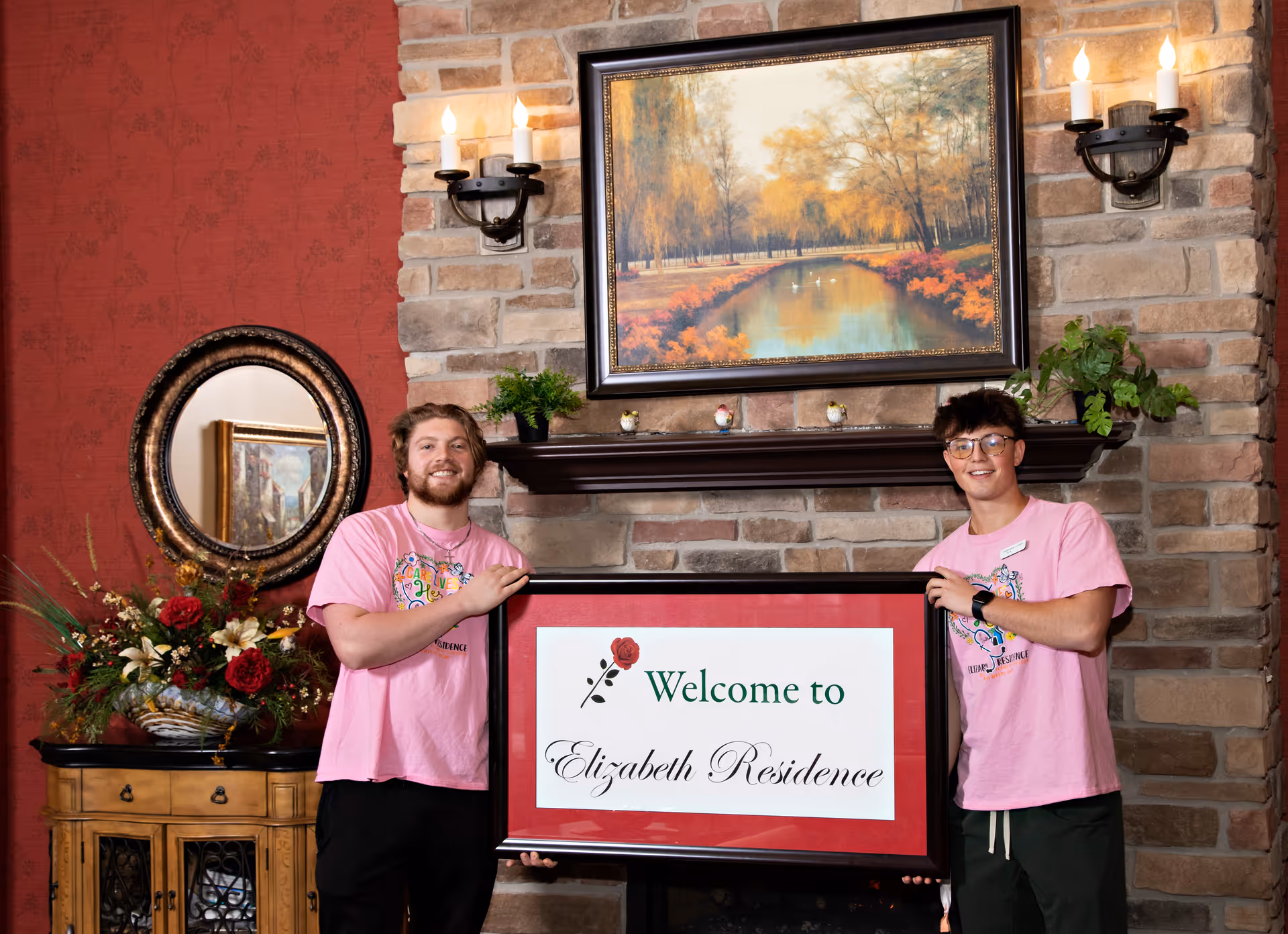 Two young men wearing pink t-shirts stand indoors in front of a stone fireplace holding a framed sign that reads 'Welcome to Elizabeth Residence'. The room has a red patterned wall, a decorative mirror, a floral arrangement on a wooden cabinet, and a framed painting of a serene autumn landscape above the fireplace.