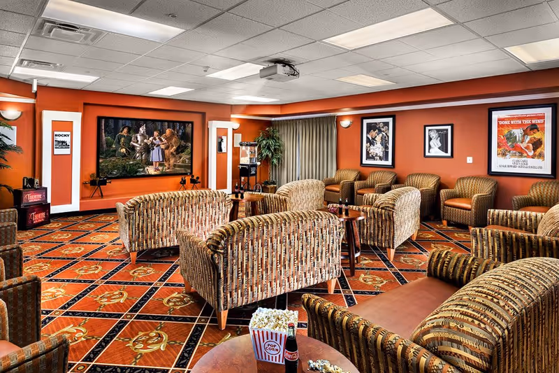 A cozy movie theater room with patterned armchairs and sofas arranged facing a large screen showing a scene from The Wizard of Oz. The room has warm orange walls decorated with framed classic movie posters including Rocky and Gone with the Wind. There is a popcorn machine in the corner and a table with popcorn and a bottle of Coca-Cola in the foreground.