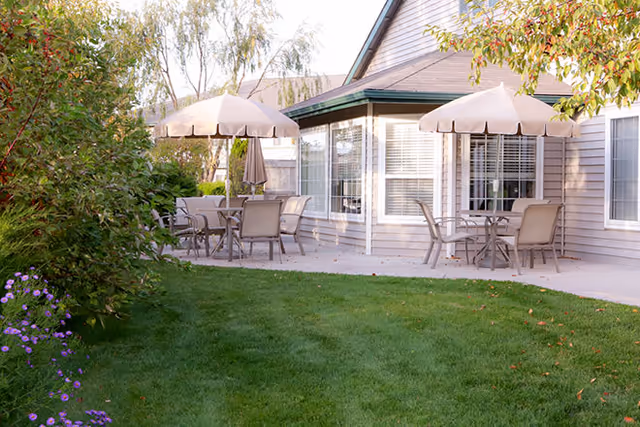 Outdoor patio with round tables, chairs and tan umbrellas on a concrete patio beside a lawn and the building exterior.