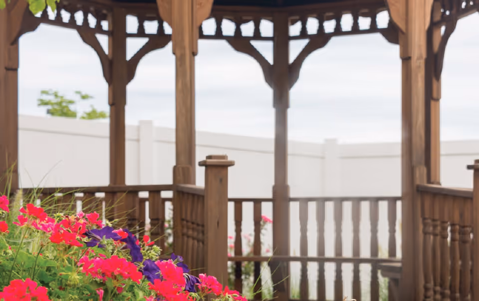 Wooden gazebo structure with decorative trim and railing, surrounded by vibrant pink and purple flowers, with a white fence in the background under a cloudy sky.