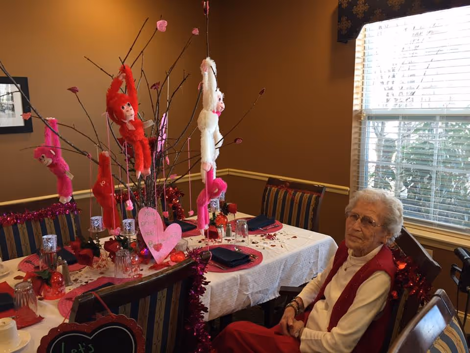 An elderly woman sitting at a dining table decorated with Valentine's Day themed decorations including pink and red plush monkeys hanging from branches, heart-shaped cutouts, and red roses. The room has brown walls and a window with blinds letting in natural light.