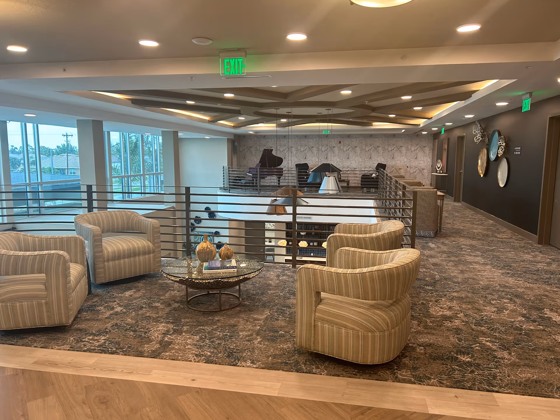 A modern indoor lounge area with four striped armchairs arranged around a round glass coffee table with decorative items on it. The space has a carpeted floor, a railing overlooking a lower level, and large windows letting in natural light. The ceiling features recessed lighting and a geometric design. In the background, there is a grand piano and additional seating along a wall with decorative art pieces.