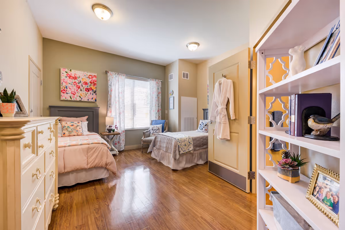 Bright, well-decorated shared bedroom with two single beds, wooden floors, dresser, shelving, and a window with floral curtains.