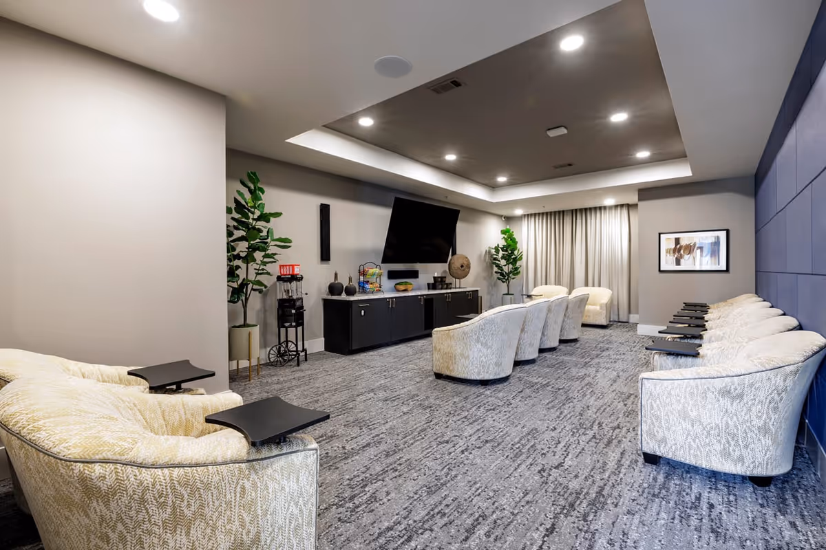 Contemporary lounge/media room with rows of upholstered chairs facing a large wall-mounted TV and console, carpeted floor and recessed lighting.
