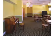 Carpeted communal room with an upright piano, round tables and chairs, a television, and ceiling fans.