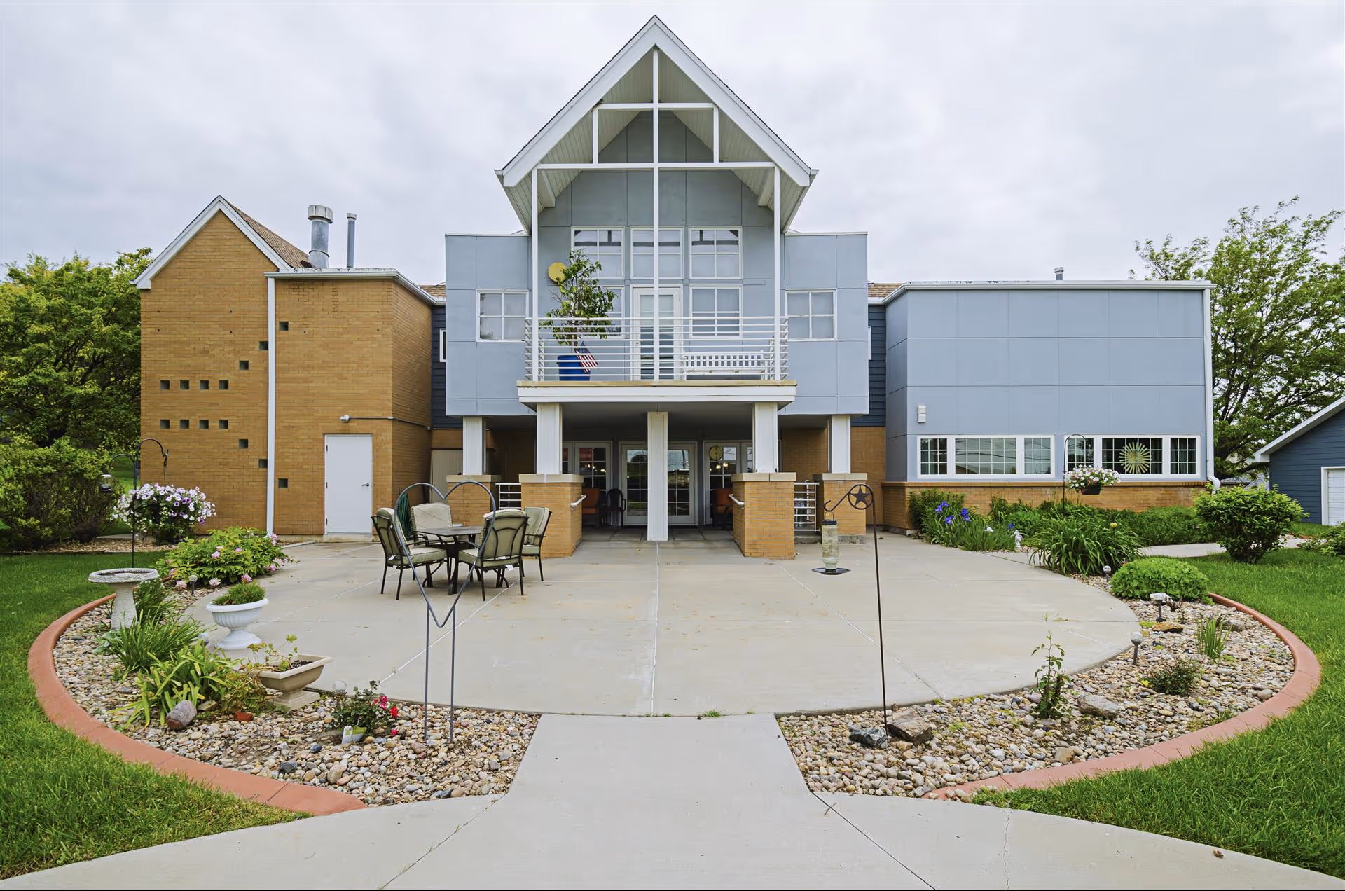 Front entrance of a two-story senior living building with a covered balcony, patio seating, and landscaped courtyard.