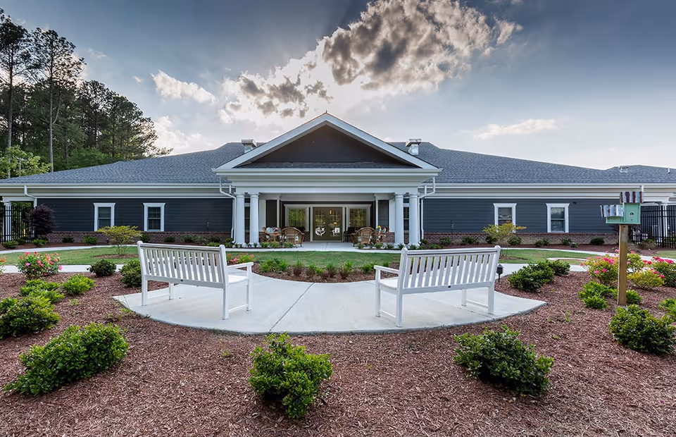 Exterior view of a single-story senior living facility building with a covered entrance supported by white columns. In front of the building, there is a landscaped garden area with mulch, green bushes, and two white benches facing each other on a concrete pathway. The sky above shows clouds with sunlight breaking through.