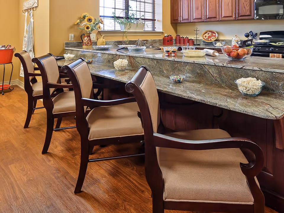A kitchen area with a granite countertop bar and four wooden chairs with beige cushions. On the counter are bowls of popcorn, candy, a bowl of apples, and various kitchen items. There is a window with blinds and a vase with sunflowers on the counter.