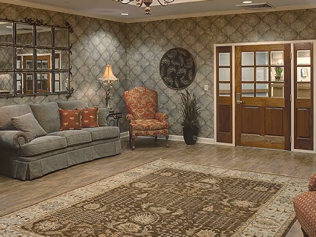 A cozy living room area with a gray sofa adorned with red patterned pillows, a red patterned armchair, a floor lamp, a large decorative mirror on the wall, a round wall art piece, a potted plant, wooden double doors with glass panels, and a large patterned area rug on a wooden floor.