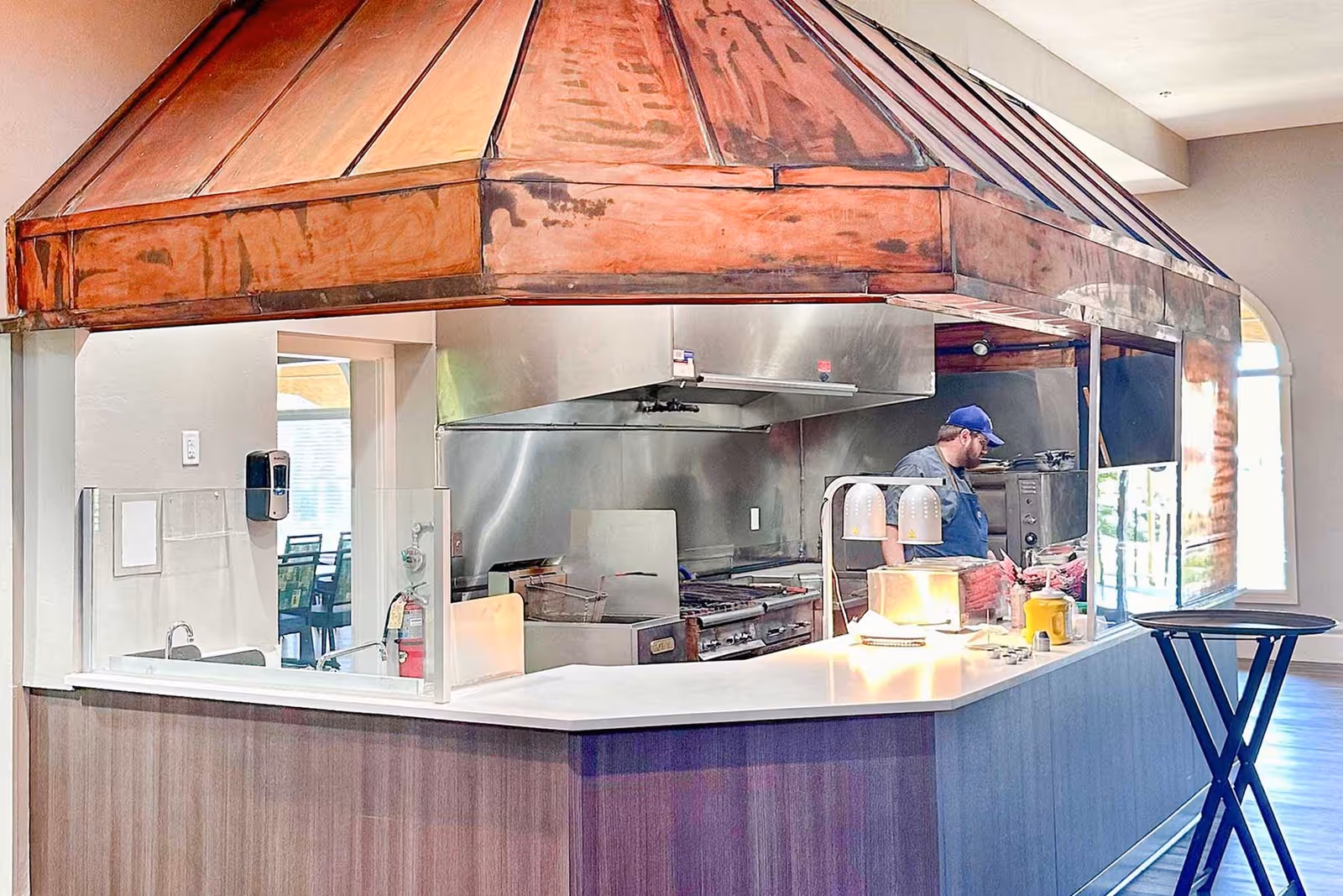 Open commercial kitchen and serving counter with a large copper hood and a staff member preparing food behind the counter.