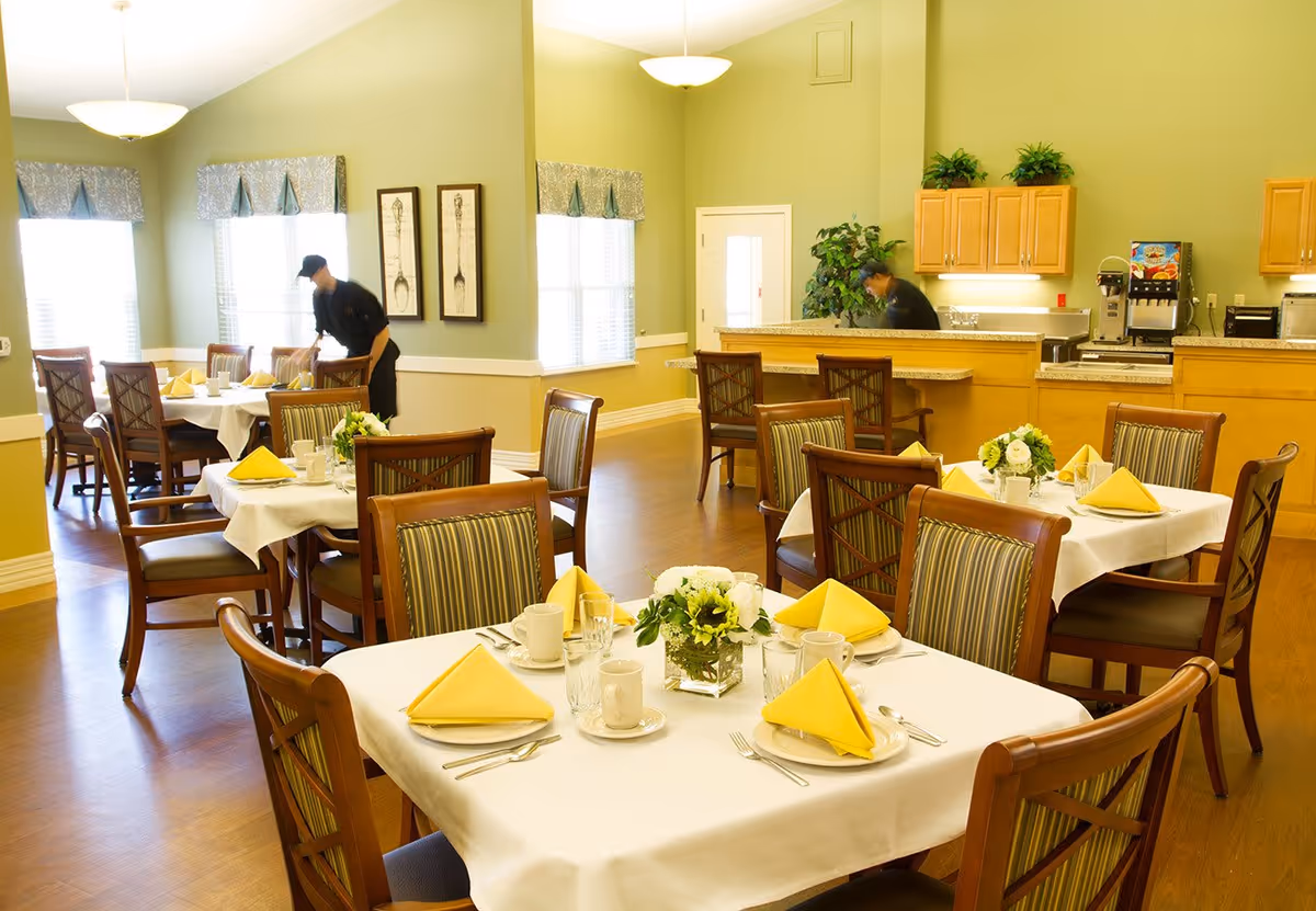A bright dining room with several tables set with white tablecloths, yellow folded napkins, plates, cups, and silverware. There are wooden chairs with striped cushions around the tables. Two staff members are seen arranging the tables and working behind a counter with wooden cabinets and a coffee machine. The walls are painted light green and there are windows with valances letting in natural light.