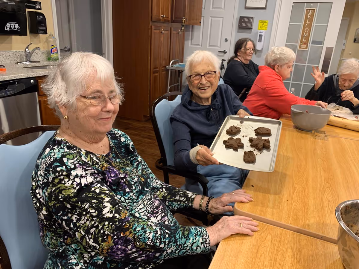 A group of elderly women sitting around a wooden table in a kitchen area. One woman is smiling and holding a baking tray with freshly baked cookies shaped like gingerbread men and other shapes. The kitchen has wooden cabinets, a sink, and a dishwasher. Other women are engaged in activities at the table.