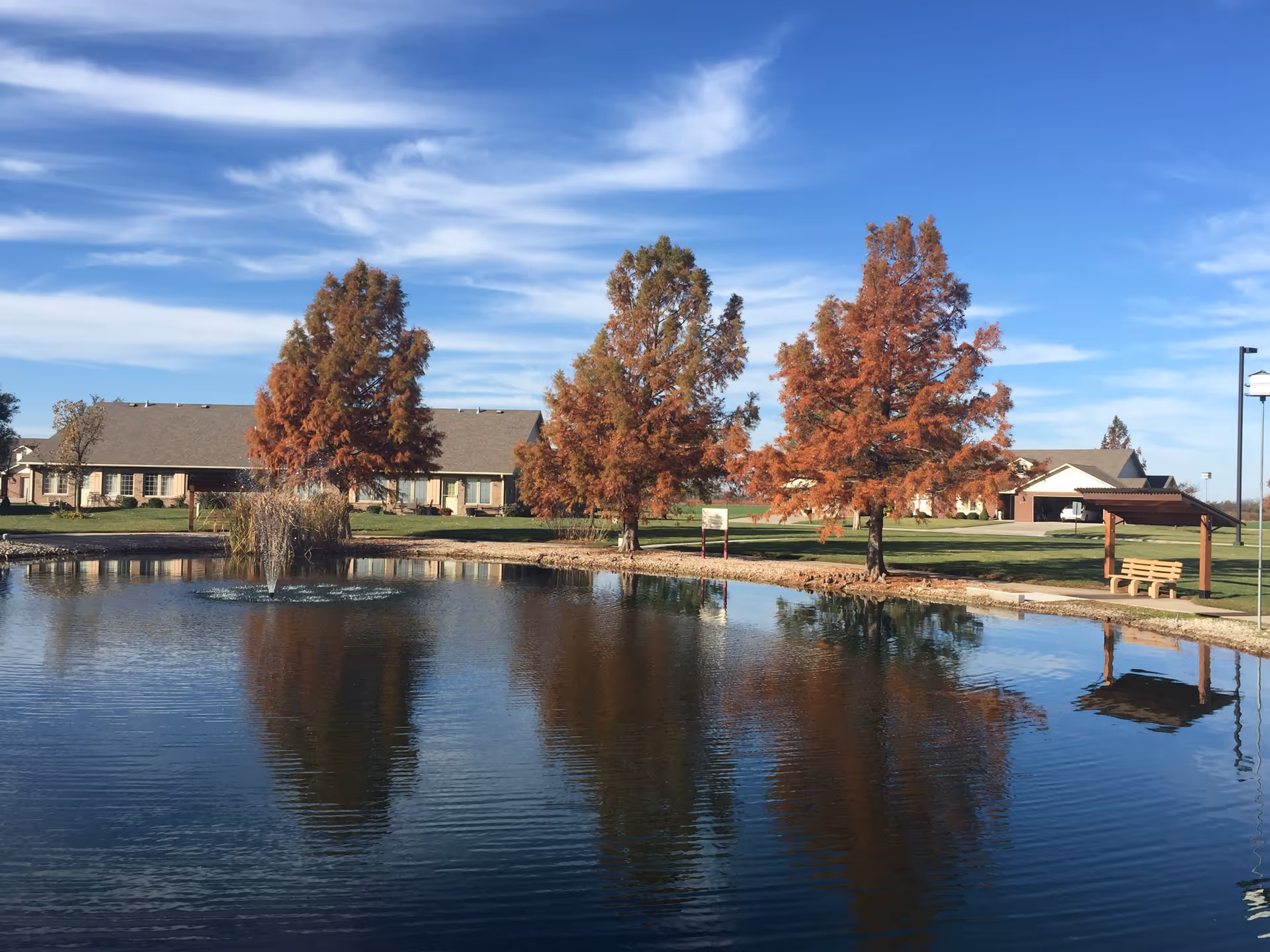 A serene outdoor scene at The Wesley featuring a pond with a small fountain in the center, surrounded by green grass and trees with autumn-colored leaves. In the background, there are single-story buildings and a wooden bench under a small pergola near the water's edge. The sky is blue with wispy clouds.