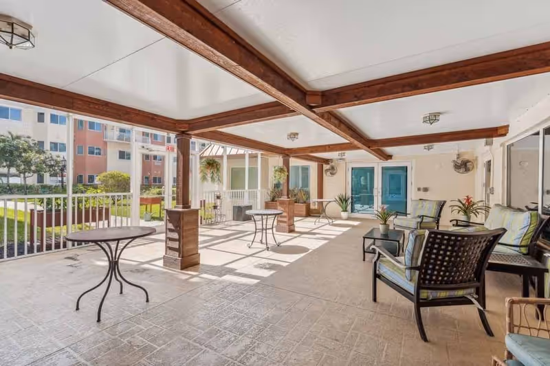 Covered outdoor patio area with wooden beams on the ceiling, several small round tables, cushioned chairs, potted plants, and a view of a grassy courtyard and multi-story building in the background.