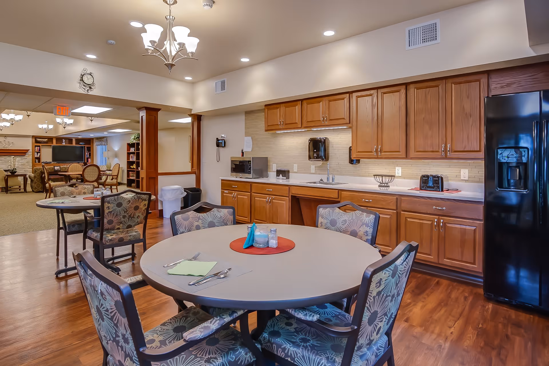 A dining area in a senior living facility featuring a round table with floral-patterned chairs, set with utensils and napkins. The background shows a kitchenette with wooden cabinets, a microwave, toaster, and a black refrigerator. Beyond the dining area, there is a lounge with more seating, a TV, and a fireplace.
