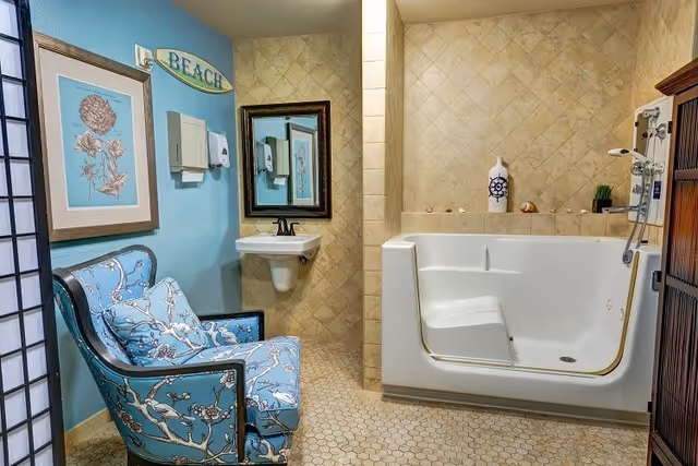 A bathroom with beige tiled walls and floor, featuring a white walk-in bathtub with a built-in seat and hand-held shower. There is a small wall-mounted sink with a mirror above it. A blue armchair with floral patterns is placed against a light blue wall decorated with framed artwork and a wooden sign that says 'BEACH'.