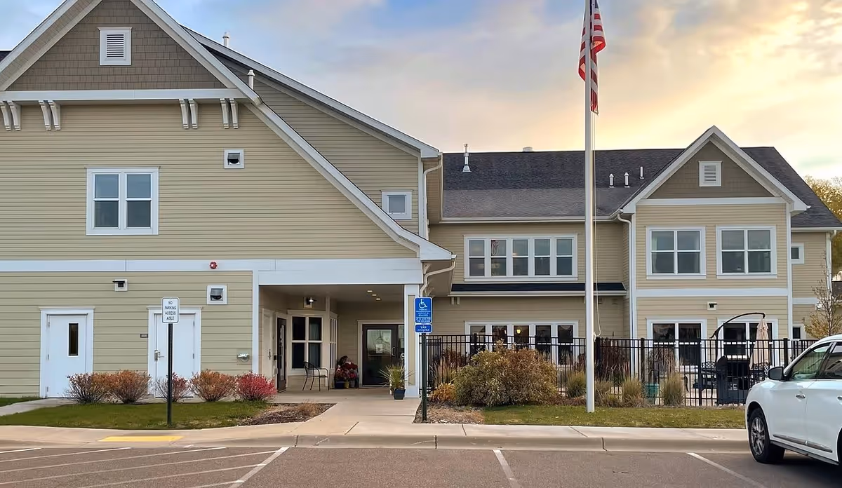 Front exterior of a two-story beige senior living building with an entrance canopy, American flag, and a parked car.