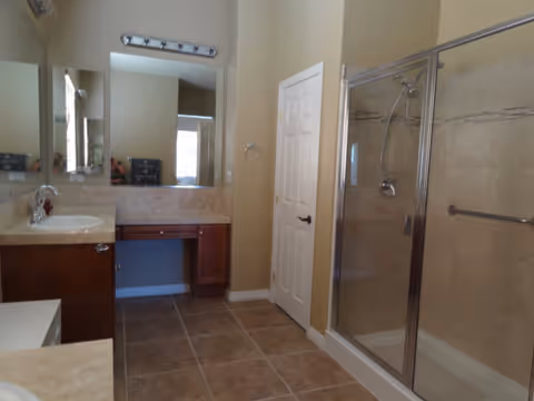 A clean bathroom featuring a glass-enclosed shower with a chrome showerhead and handle, a vanity with a sink and faucet, a large mirror above the vanity, tiled floor, and a white door. The walls are painted beige and there is a towel ring mounted near the door.