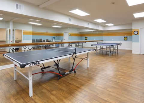 Indoor recreation room with two ping-pong tables on a wood floor, mirrored wall, and stacked chairs along the side.