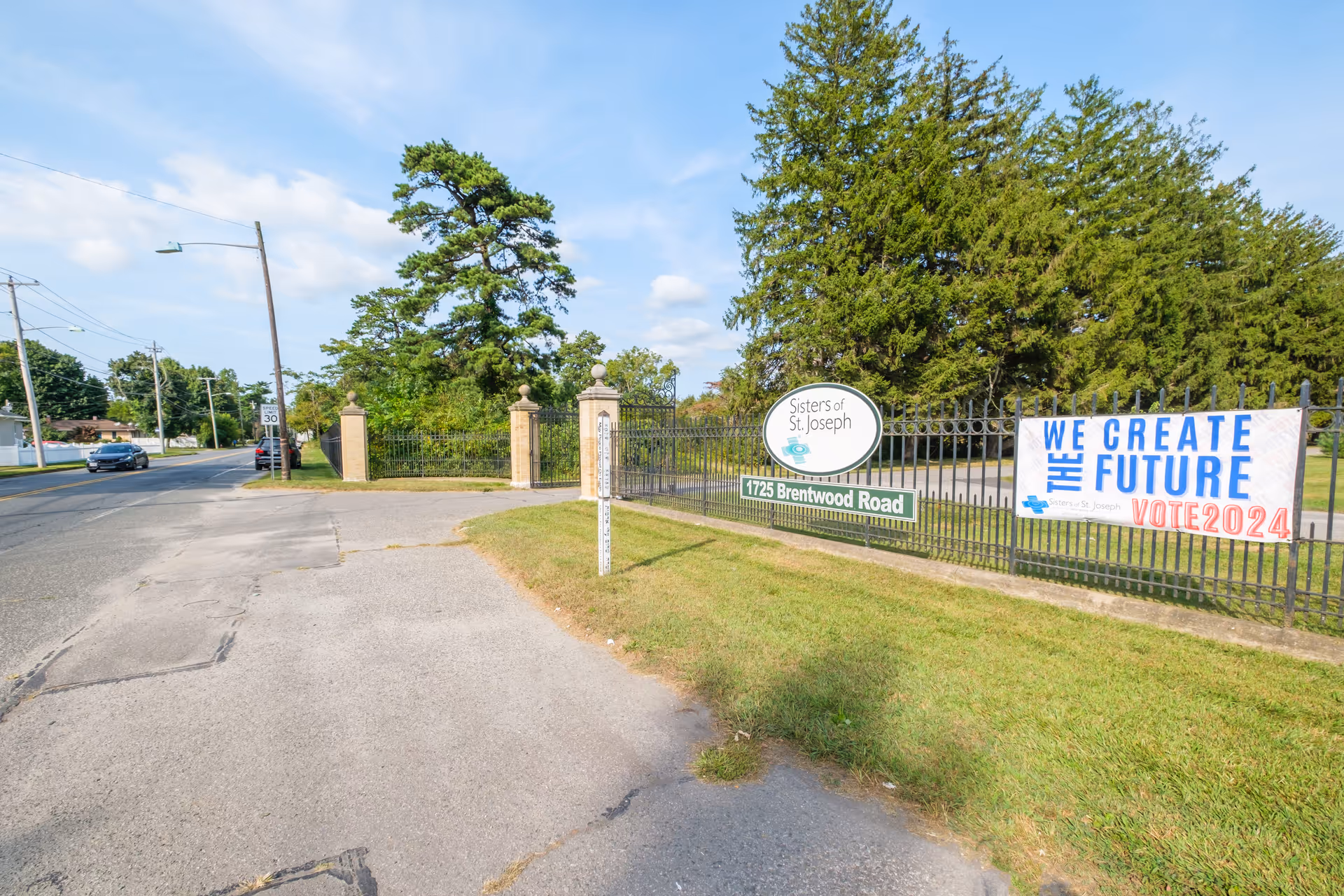 Entrance to Sisters of St. Joseph at 1725 Brentwood Road with a black iron fence and stone pillars, a banner reading 'We Create The Future Vote 2024', a speed limit sign on the road, and trees in the background under a partly cloudy sky.