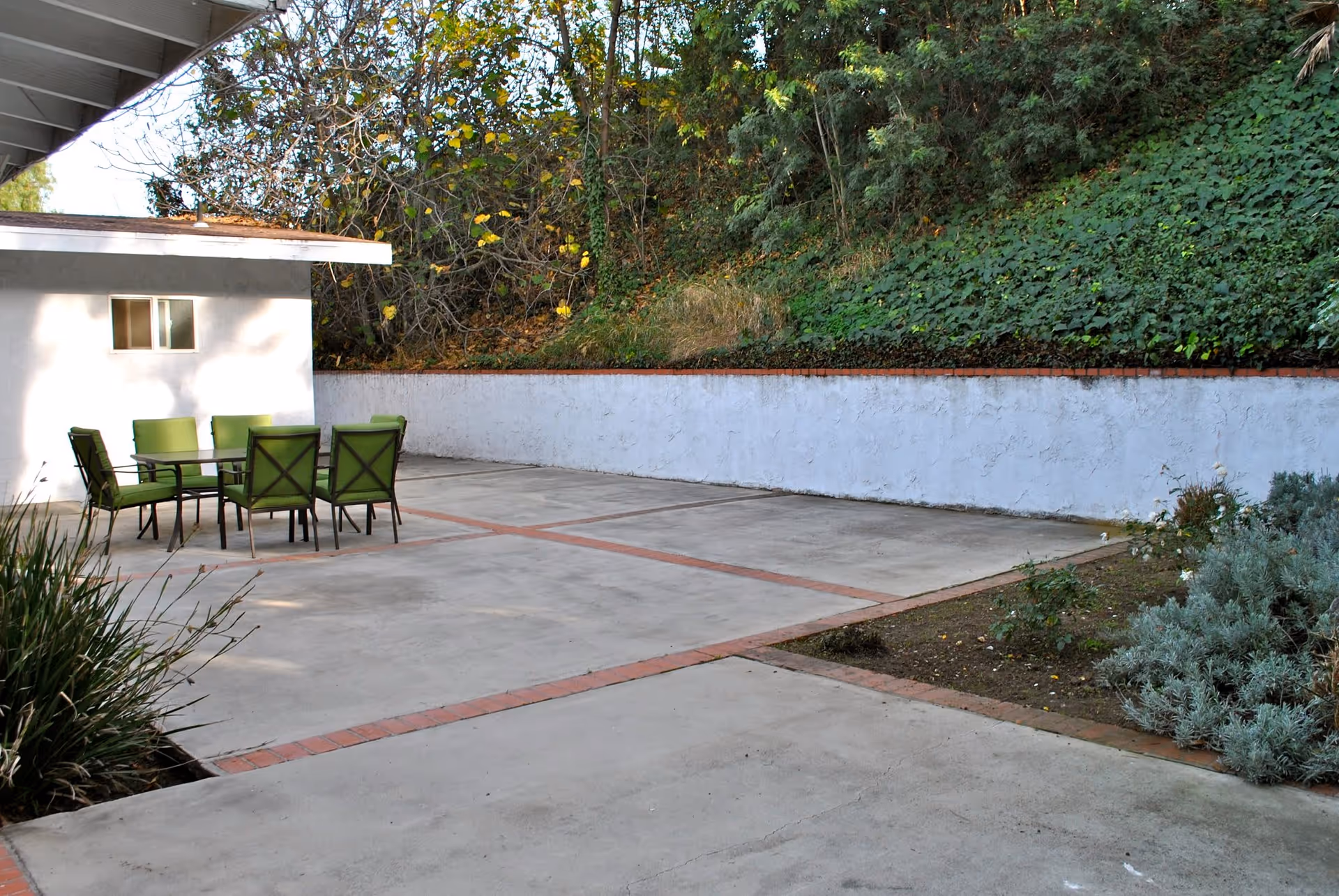 Outdoor patio area with a concrete floor and a table surrounded by six green cushioned chairs. The patio is adjacent to a white building wall and bordered by a white retaining wall with greenery and trees behind it.