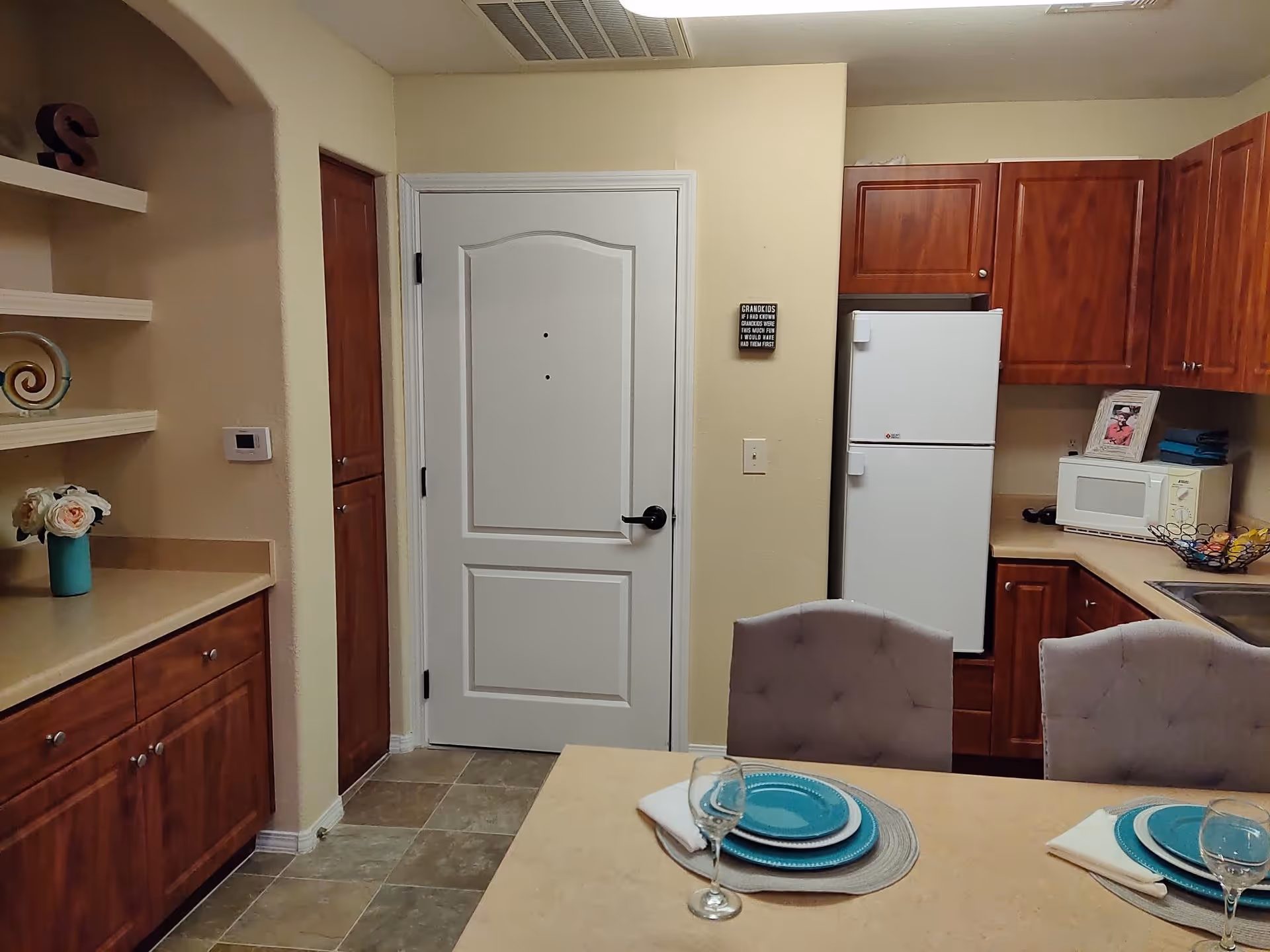 Interior view of a kitchen area with wooden cabinets, a white refrigerator, a microwave, and a sink. There is a countertop with two place settings including plates, napkins, and wine glasses. A white door is visible in the background, along with some decorative items on shelves to the left.
