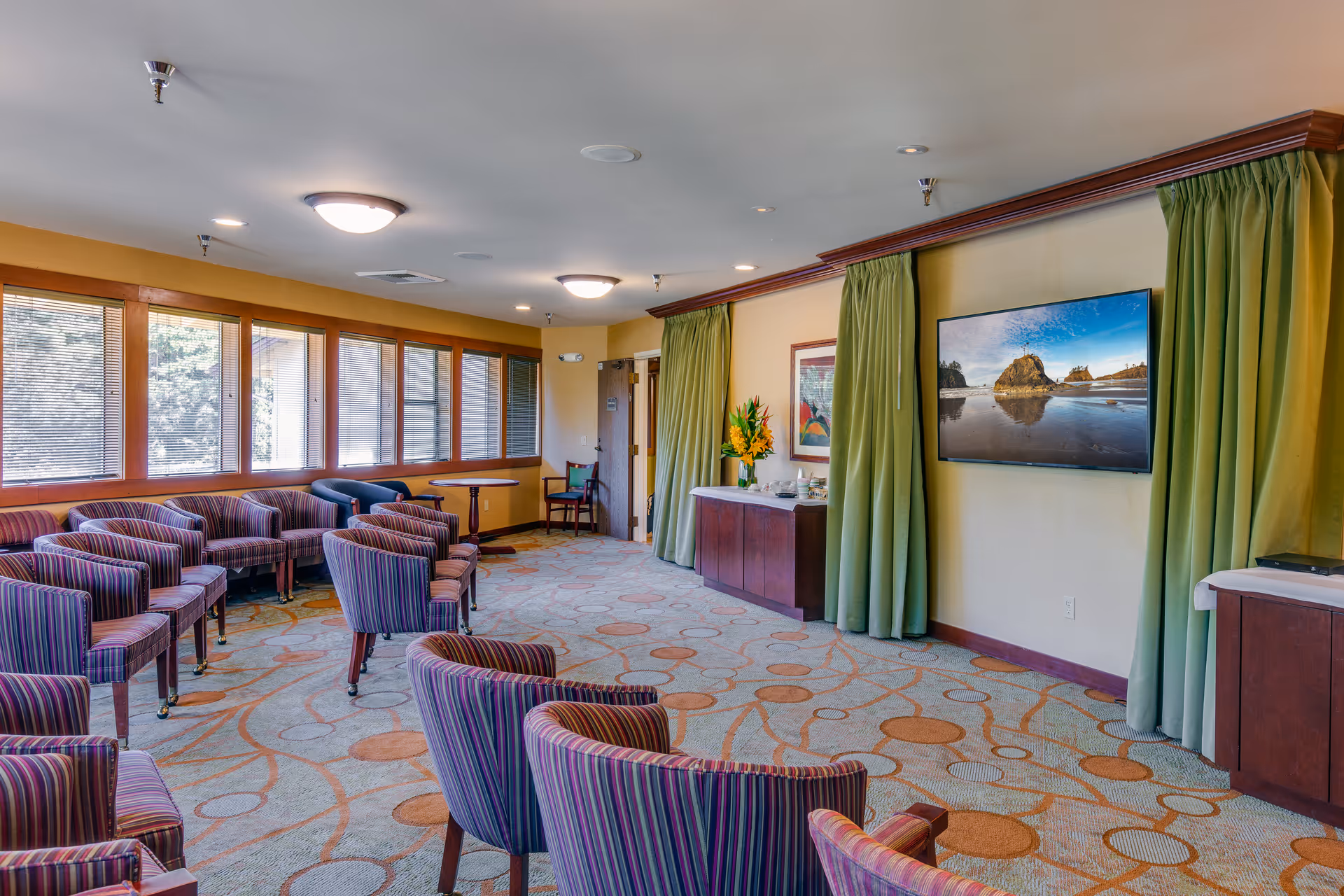 A spacious common area with multiple striped upholstered chairs arranged in rows facing a wall-mounted flat screen TV displaying a scenic beach image. The room has large windows with blinds on one side, green curtains framing the TV, a carpet with circular patterns, and wooden furniture including a cabinet with a flower arrangement and a small table with chairs near the door.