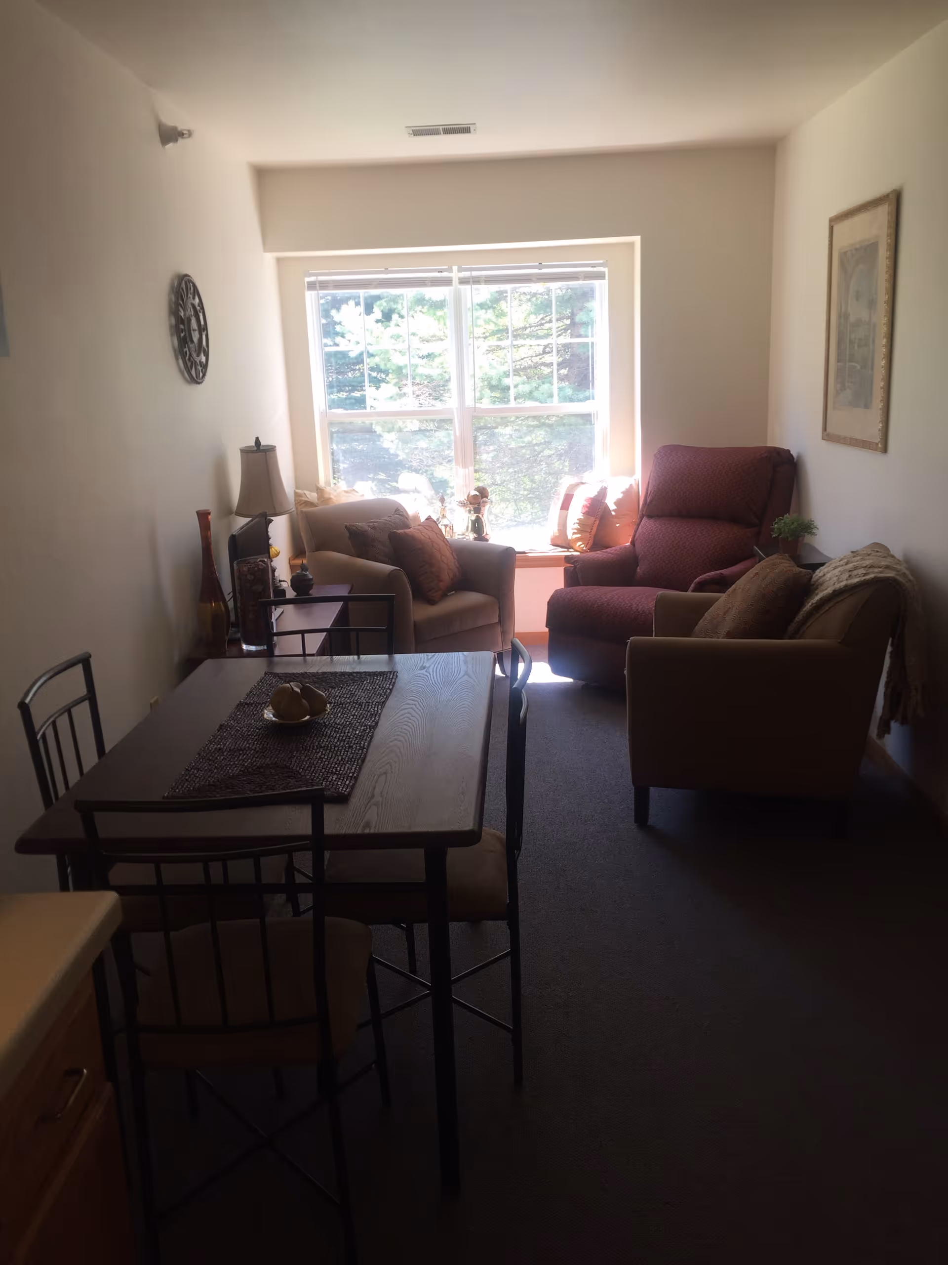 A cozy living room area with a large window letting in natural light. The room features a red armchair, two beige armchairs with brown cushions, a small side table with a lamp, and a dining table with four chairs. There is a decorative clock on the wall and a framed picture on the opposite wall.