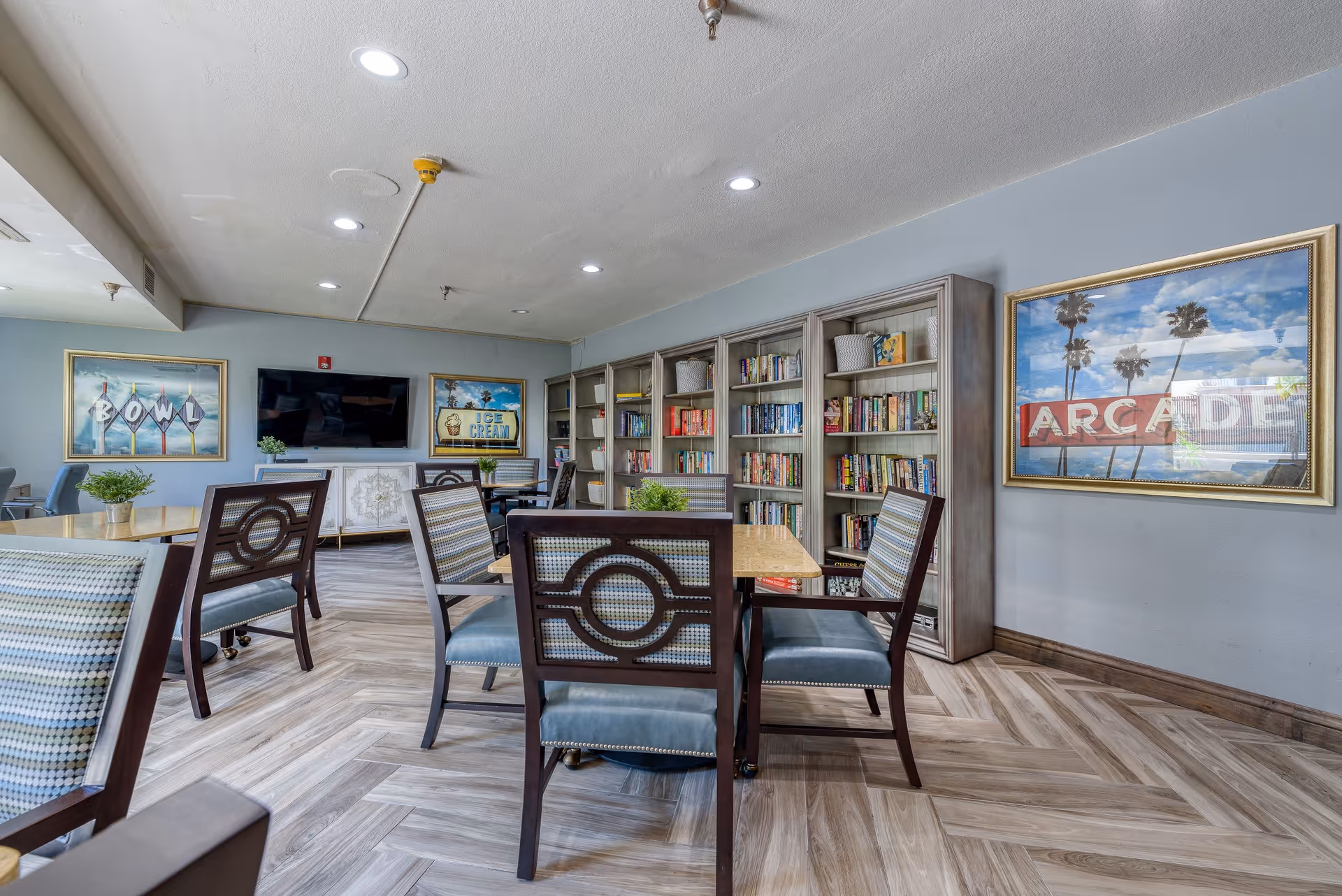 A senior living facility common area with several tables and chairs arranged for seating. There are bookshelves filled with books along one wall, a flat-screen TV mounted on another wall, and framed vintage-style signs that read 'BOWL', 'ICE CREAM', and 'ARCADE'. The room has wood-patterned flooring and soft blue-gray walls.