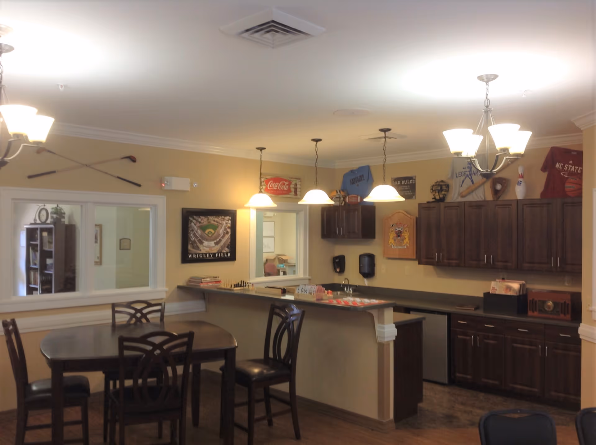 Communal dining area with a table and chairs, a serving counter and small kitchen with dark wood cabinets and pendant lights.