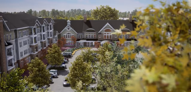Aerial view of Broadview Senior Living at Purchase College showing a multi-story building with a pitched roof surrounded by trees and a parking lot with several cars. The building has multiple windows and balconies, and the scene is set on a clear day with a blue sky.