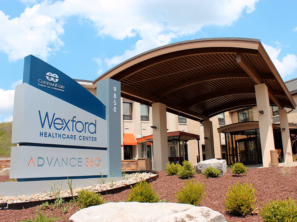 Entrance of Wexford Healthcare Center showing a large covered drop-off area with pillars, landscaping with bushes and rocks, and a sign displaying the facility's name and address under a partly cloudy sky.